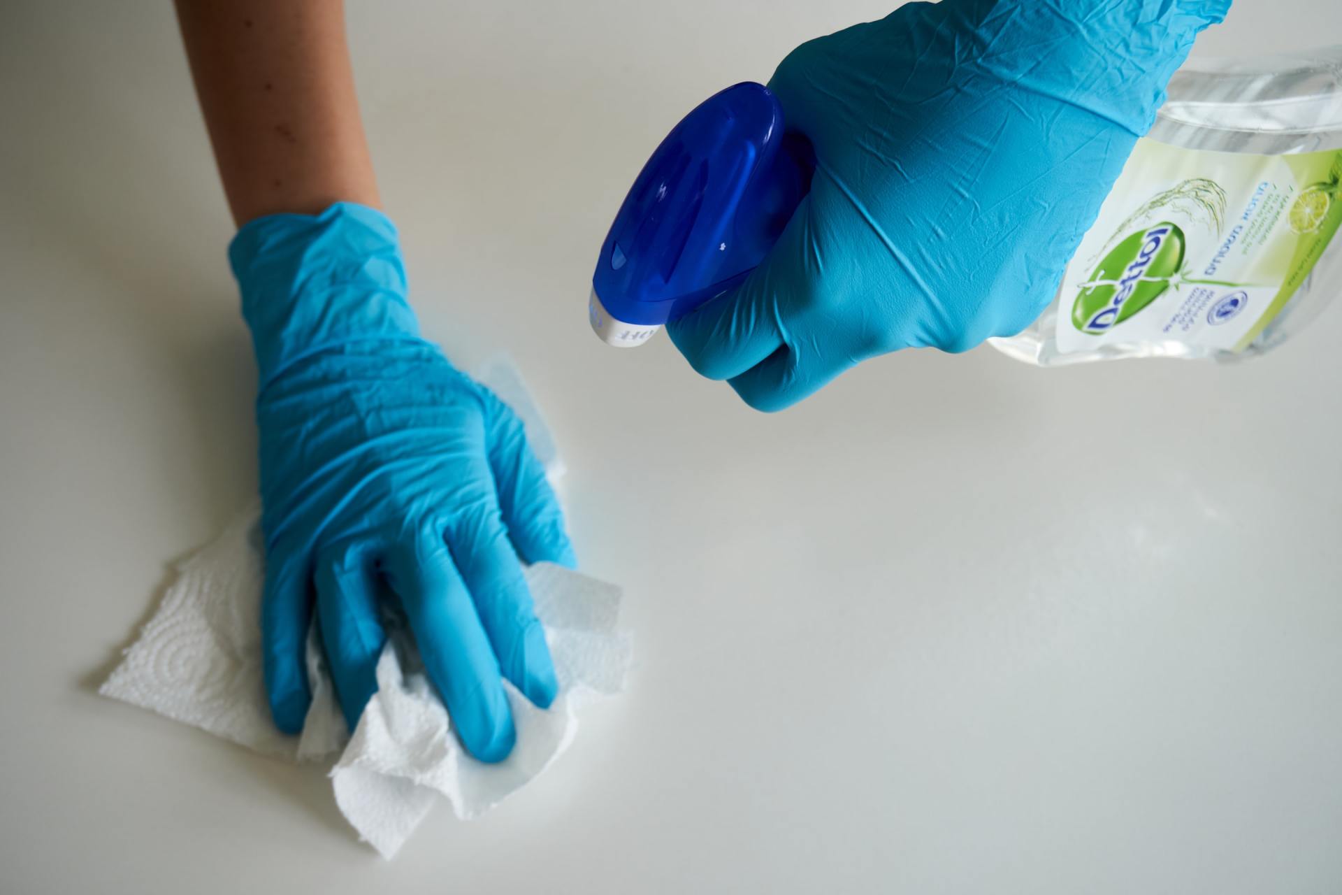 Hands in blue gloves spraying cleaner on a surface and wiping it with a white paper towel.