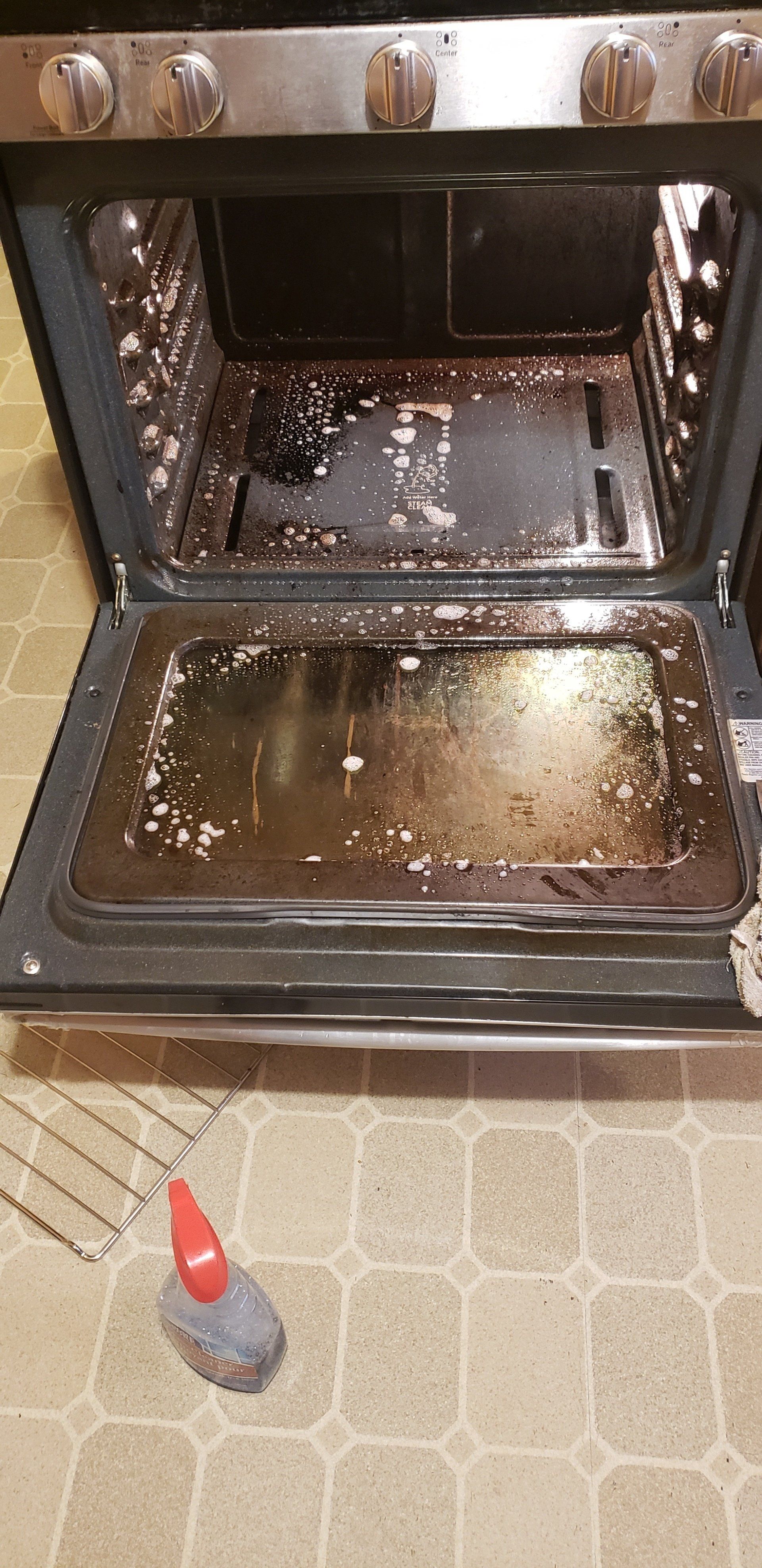 An open oven with a dirty interior and a cleaning sponge on the floor.